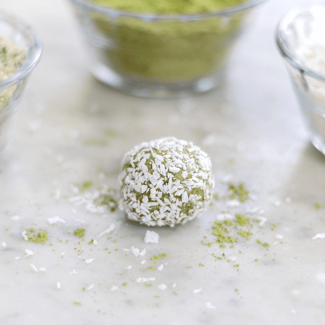 close up of the Matcha Coconut fat bomb: round ball covered in coconut on a marble countertop with glass bowls of ingredients in the background