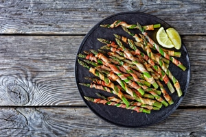 Bacon wrapped asparagus easy side dish grilled with garlic and sea salt served on a black plate lemon on an old wooden background