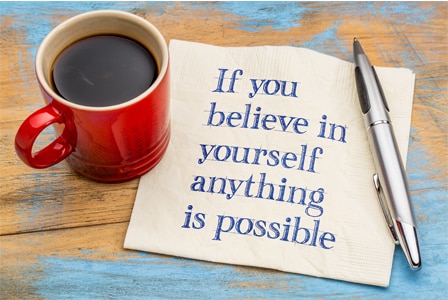 a red coffee mug full of black coffee with a paper napkin with blue words, "If you believe in yourself anything is possible" and a pen is next to the napkin